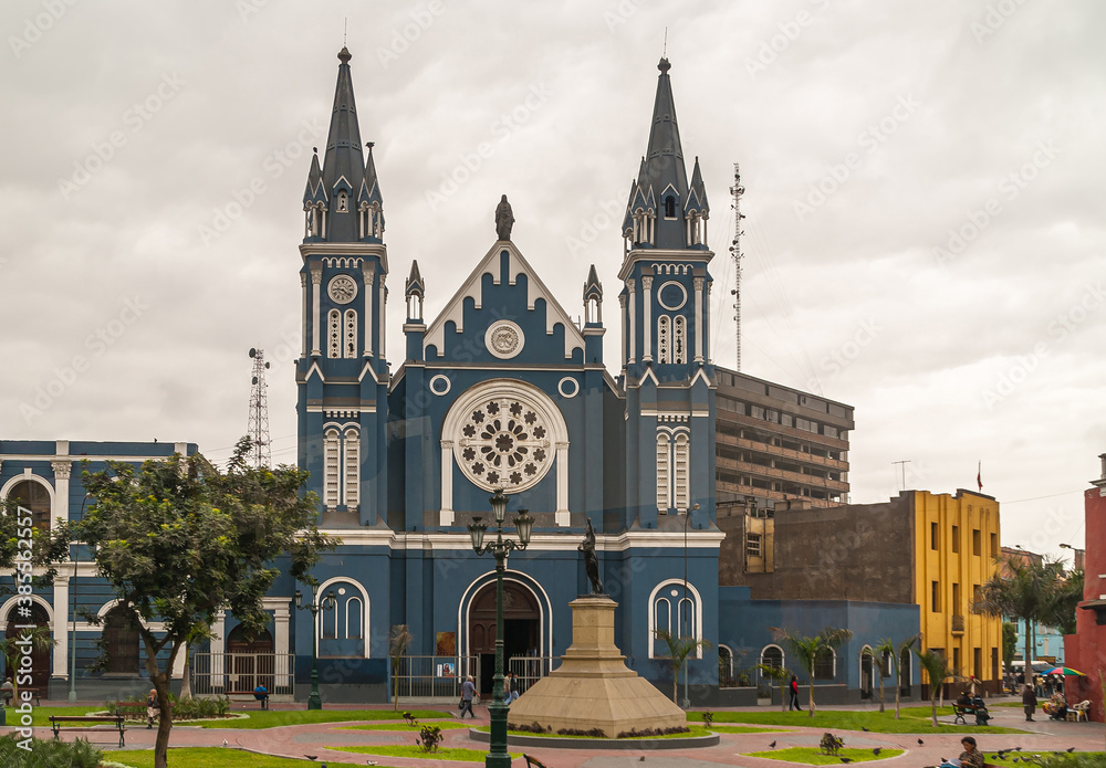 Lima, Peru - December 4, 2008: Blue facade with white trim of La Recoleta Church under silver-brown cloudscape with green foliage in front. Housing around. Statue of Freedom.