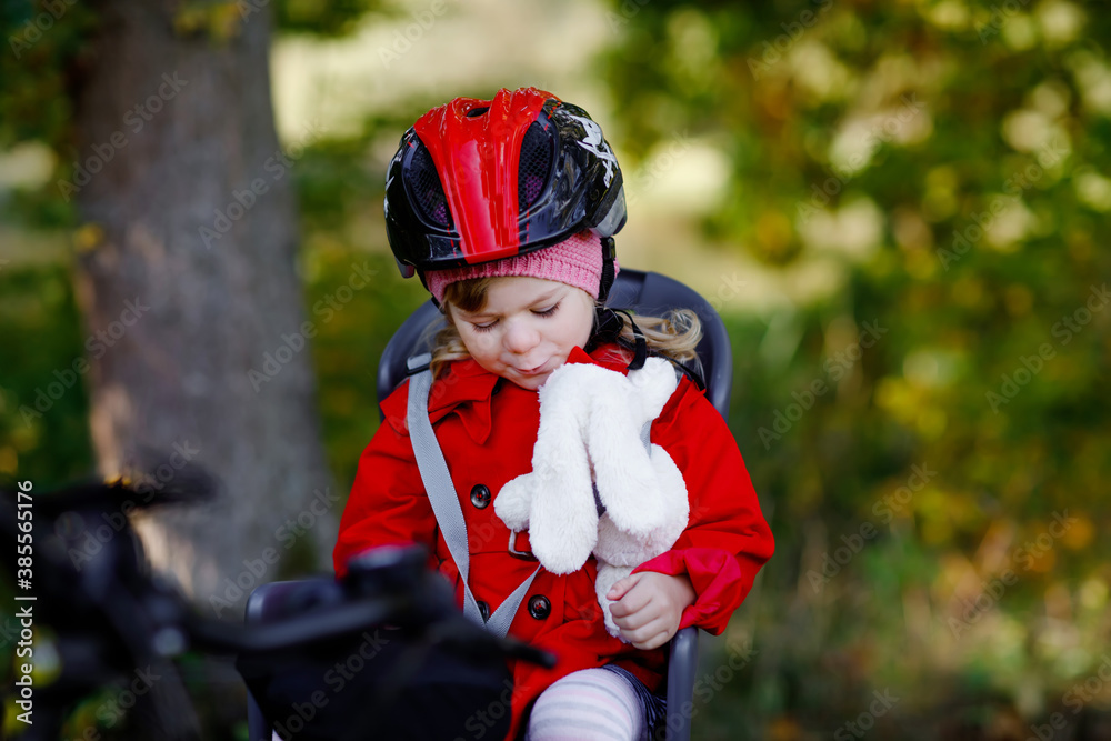 Little toddler girl with security helmet on head sitting in bike seat of her mother or father bicycle. Safe and child protection concept. Family ctivity trip on autumn day. Kid holding push soft toy Little toddler girl with security helmet on head sitting in bike seat of her mother or father bicycle. Safe and child protection concept. Family ctivity trip on autumn day. Kid holding push soft toy