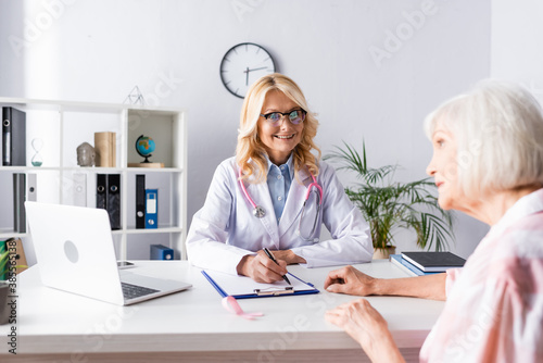 Selective focus of doctor writing on clipboard and looking at patient in clinic