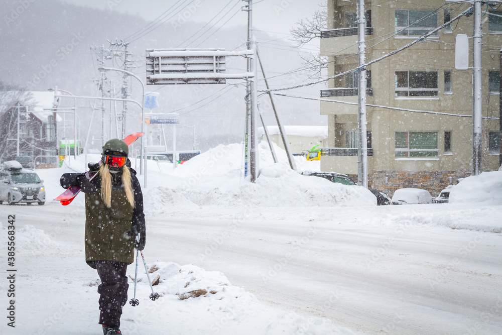 Fototapeta premium Picture of ski resort town while snowing and the street is covered with snow and a woman wear dark green jacket carring ski walking on the sidewalk.