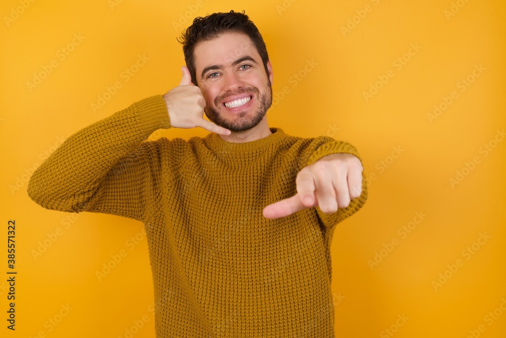 Young man wearing casual sweater and over isolated yellow background smiling cheerfully and pointing to camera while making a call you later gesture, talking on phone Young man wearing casual sweater and over isolated yellow background smiling cheerfully and pointing to camera while making a call you later gesture, talking on phone
