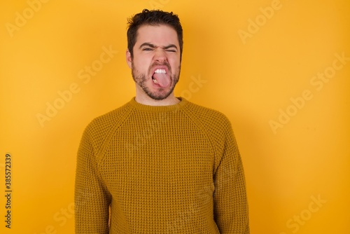 Young man wearing casual sweater and over isolated yellow background sticking tongue out happy with funny expression. Emotion concept. © Roquillo