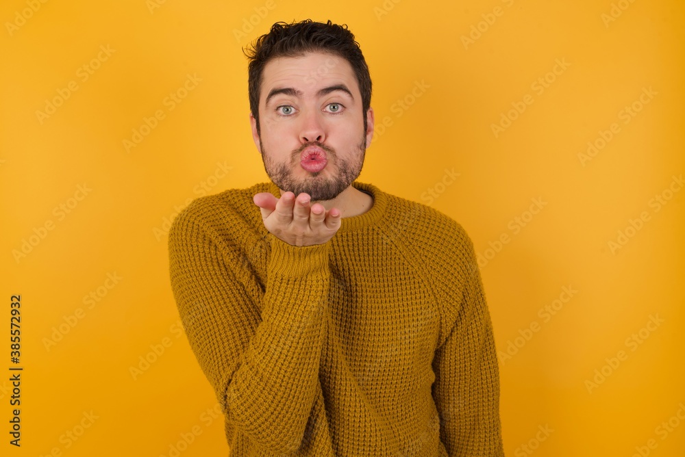 Young man wearing casual sweater and over isolated yellow background looking at the camera blowing a kiss with hand on air being lovely and sexy. Love expression. Young man wearing casual sweater and over isolated yellow background looking at the camera blowing a kiss with hand on air being lovely and sexy. Love expression.