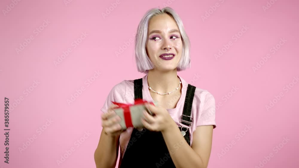 Cute young woman with colorful hair holding gift box with bow, she wonders what is inside. Pink wall background. Girl smiling, she is happy to get present. Cute young woman with colorful hair holding gift box with bow, she wonders what is inside. Pink wall background. Girl smiling, she is happy to get present.