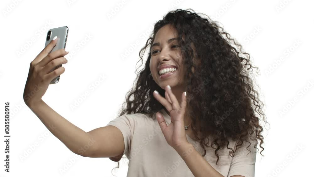 Close-up of cute mexican girl with dark curly hairstyle, saying hello and smiling, having video call and waving hand at front camera, standing in t-shirt over white background Close-up of cute mexican girl with dark curly hairstyle, saying hello and smiling, having video call and waving hand at front camera, standing in t-shirt over white background