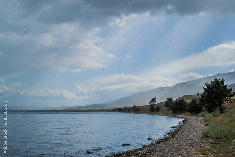 Pebbles shore with trees of clear calm undulating blue Lake Baikal, mountains on the horizon, clouds Pebbles shore with trees of clear calm undulating blue Lake Baikal, mountains on the horizon, clouds