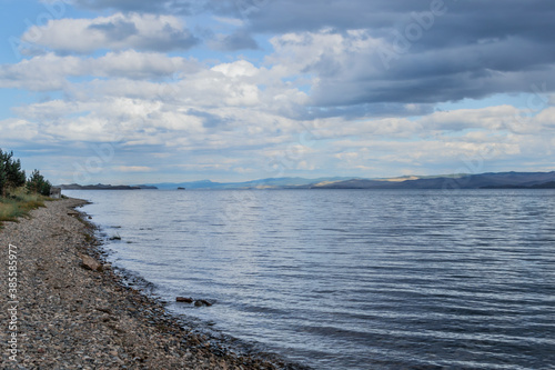 Fototapeta Naklejka Na Ścianę i Meble -  Pebbles shore of clear calm undulating blue Lake Baikal, mountains on the horizon, clouds
