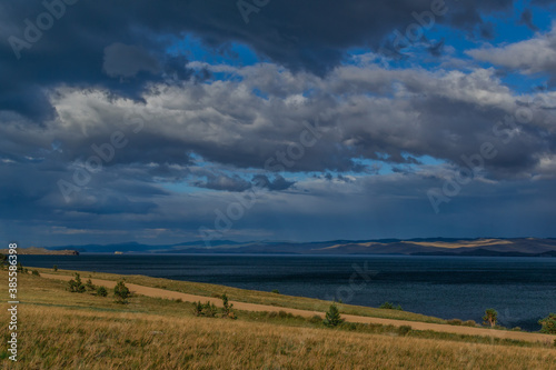 Coniferous green trees and road in yellow grassy coast of blue lake Baikal with mountains. Summer landscape in sunset light