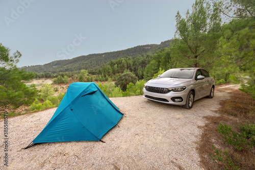 The blue tent is set up on a flat area in the forest near a car. Road trips and camping in the woods