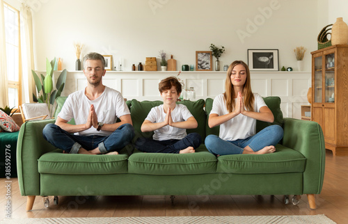 Canvas Print Serene couple and little son sitting on sofa in lotus position with closed eyes doing namaste gesture meditating in living room