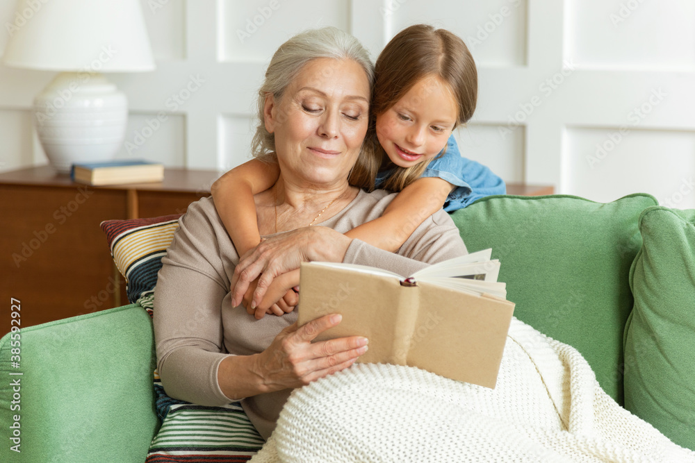 Pretty grandmother and little granddaughter reading book, sitting on comfortable couch in living room. Smiling elderly woman teaching girl child on weekend at home, children education concept. Pretty grandmother and little granddaughter reading book, sitting on comfortable couch in living room. Smiling elderly woman teaching girl child on weekend at home, children education concept.