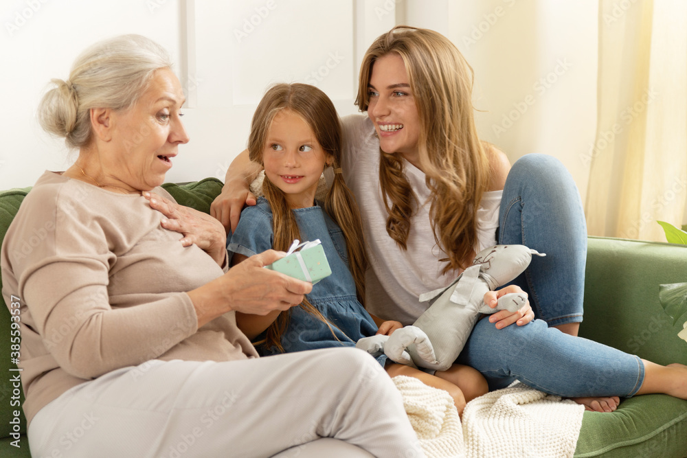 Happy shocked mature grandmother holds gift box with present from grandchild girl and smiling daughter. Three female generations together. Cute granddaughter and her mother congratulating grandma. Happy shocked mature grandmother holds gift box with present from grandchild girl and smiling daughter. Three female generations together. Cute granddaughter and her mother congratulating grandma.