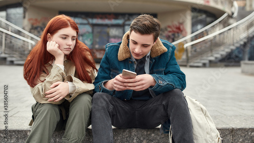 Two teenagers spending time together, sitting on the steps outdoors. Upset girl looking at her boyfriend while he is ignoring her, using his smartphone