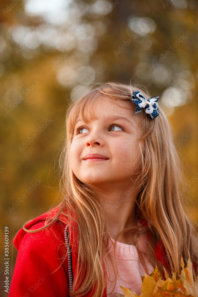 Fototapeta premium Outdoor portrait of happy blonde child girl in a red jacket holding yellow leaves. Little girl walking in the autumn park or forest. Warm sunny weather. Fall concept