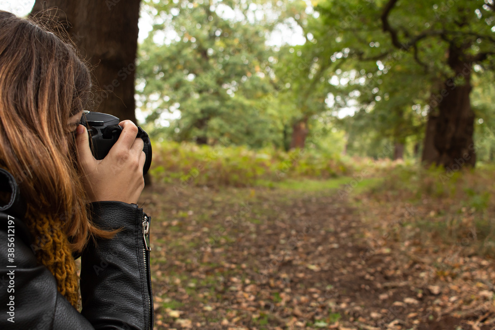 Attractive young female with a black leather jacket taking a photo with her reflex camera while walking on the park during autumn