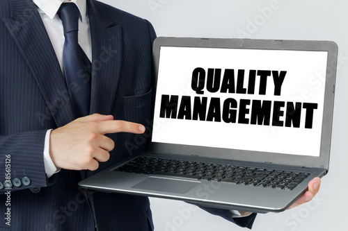 A businessman holds a laptop in his hands and shows on the screen with the text - QUALITY MANAGEMENT