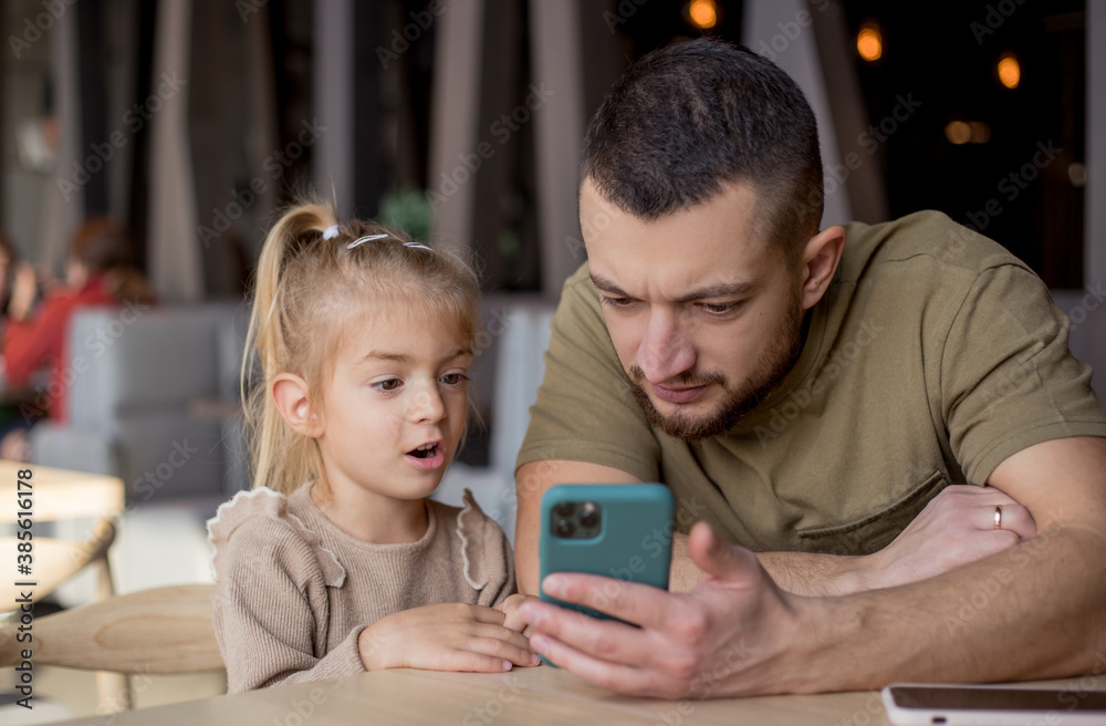 Young daddy embracing child daughter, showing funny videos on smartphone. Overjoyed little girl looking at mobile screen with father, technology addiction. Young daddy embracing child daughter, showing funny videos on smartphone. Overjoyed little girl looking at mobile screen with father, technology addiction.