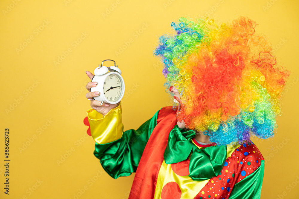 Fototapeta premium Clown standing over yellow insolated yellow background surprised holding a clock