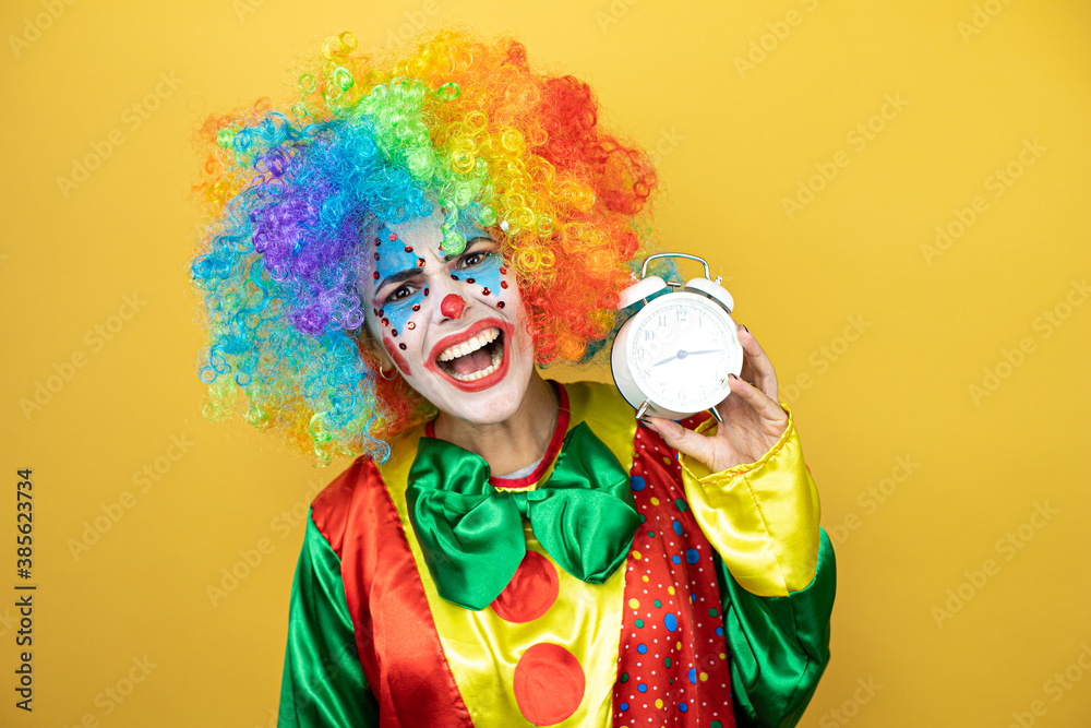 Clown standing over yellow insolated yellow background surprised holding a clock