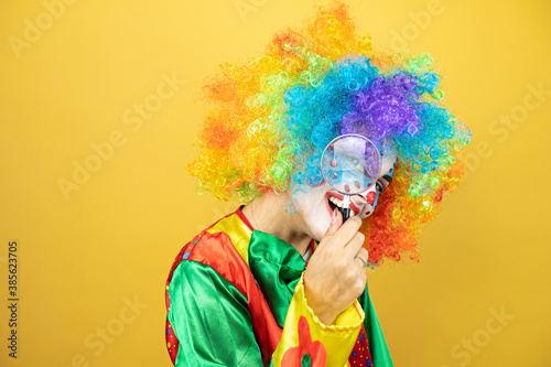 Clown standing over yellow insolated yellow background looking through a magnifying glass