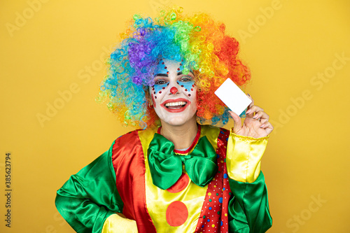 Clown standing over yellow insolated yellow background smiling and holding white card