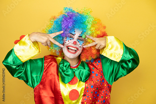 Clown standing over yellow insolated yellow background Doing peace symbol with fingers over face  smiling cheerful showing victory