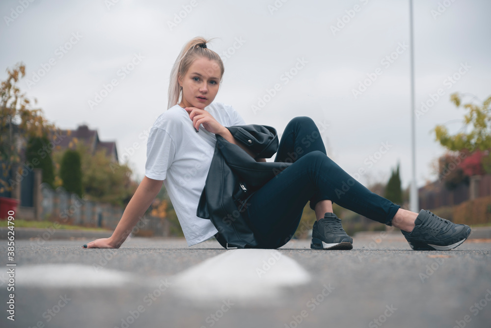 Young fresh cheerful hipster young woman sitting on the road in countryside wearing stylish summer clothes. Bright bag pack. Warm evening colors. Sunset