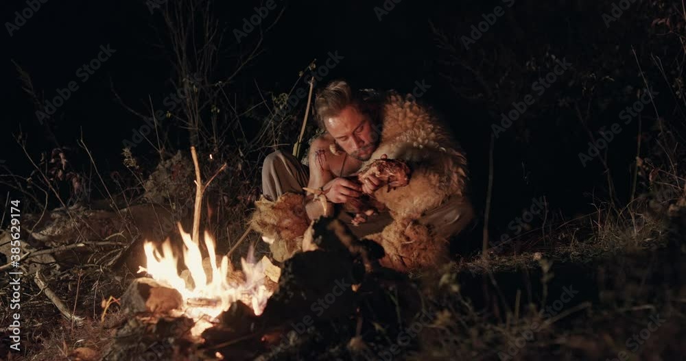 Hungry wild man eating near campfire. Low angle of hungry male barbarian eating meat while sitting near fire at night in countryside