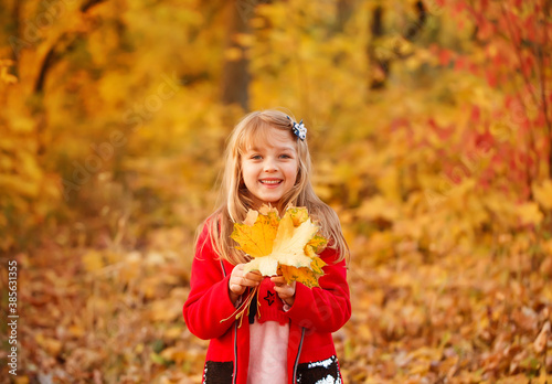 Outdoor portrait of happy blonde child girl in a red jacket holding yellow leaves. Little girl walking in the autumn park or forest. Warm sunny weather. Fall concept