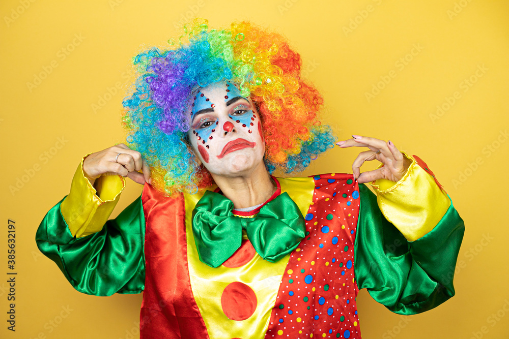 Clown standing over yellow insolated yellow background holding her t-shirt with a successful expression Clown standing over yellow insolated yellow background holding her t-shirt with a successful expression