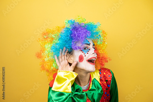 Clown standing over yellow insolated yellow background surprised with hand over ear listening an hearing to rumor or gossip
