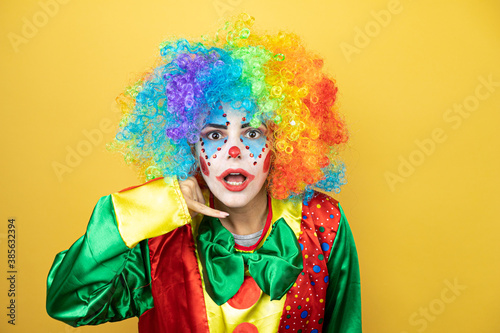 Clown standing over yellow insolated yellow background confused doing phone gesture with hand and fingers like talking on the telephone