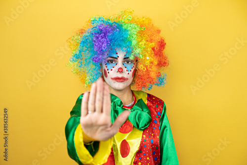 Clown standing over yellow insolated yellow background serious and doing stop sing with palm of the hand.