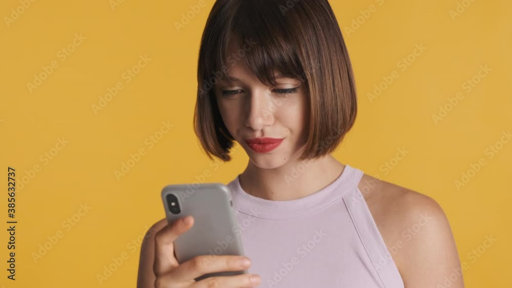 Pretty brunette girl chatting with friends online on smartphone isolated on yellow background Pretty brunette girl chatting with friends online on smartphone isolated on yellow background