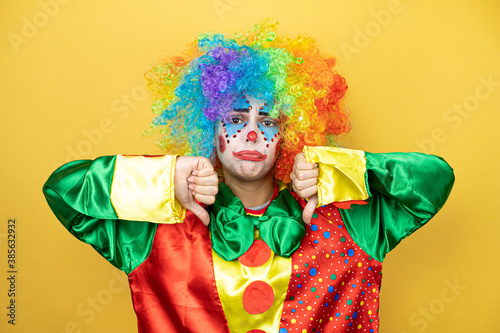 Clown standing over yellow insolated yellow background with angry face, negative sign showing dislike with thumb down