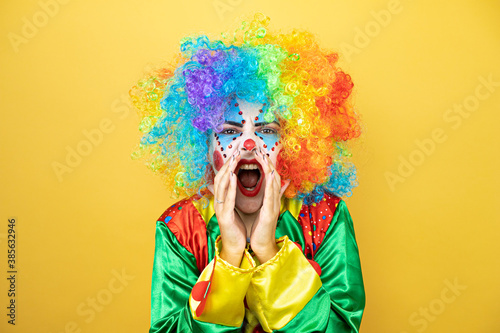 Clown standing over yellow insolated yellow background shouting and screaming loud to side with hands on mouth