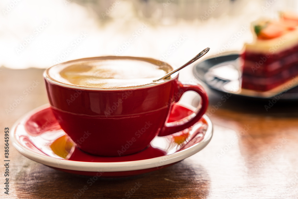 Close up of a red cup of coffee on a wooden table. In the background is a black plate with strawberry cake Close up of a red cup of coffee on a wooden table. In the background is a black plate with strawberry cake