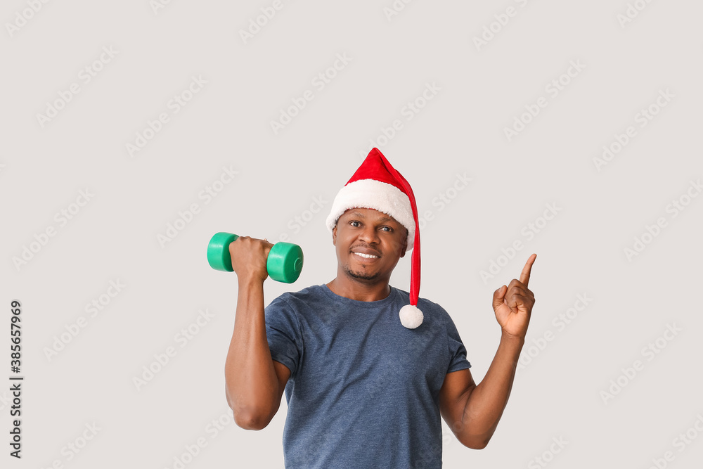 African-American man in Santa hat and with dumbbell on light background