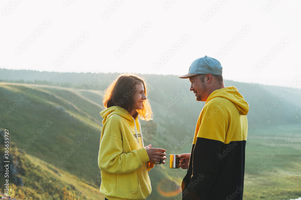 Young happy family couple travelers in casual outfits with tea in morning on beautiful view background. Local tourism, the weekend trip Young happy family couple travelers in casual outfits with tea in morning on beautiful view background. Local tourism, the weekend trip