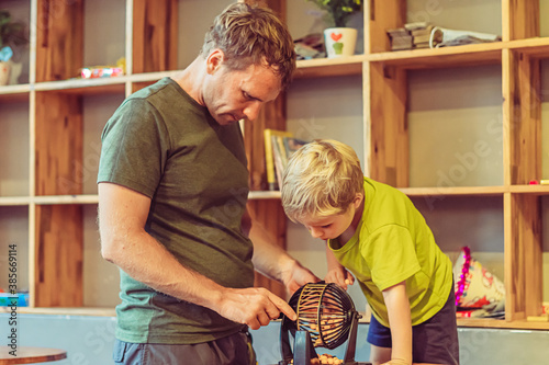 Father blond son boy play lotto, round sphere spins with small orange balls numbers written, table loto game. Children home education learn numeros find common language develop logical thinking