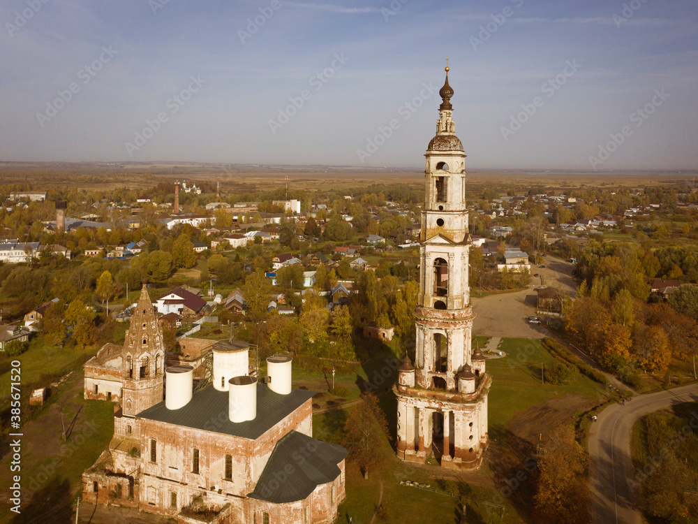 Aerial shot of ruins of a Russian church and tall bell tower standing in a small town. Autumn colours, long shadows, blue sky