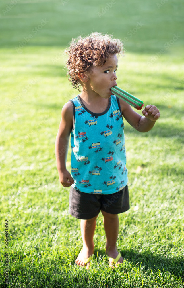 Cute African American Boy eating a giant popsicle outdoors in the summertime. He takes a big bite of a cool snack on a warm summer day Cute African American Boy eating a giant popsicle outdoors in the summertime. He takes a big bite of a cool snack on a warm summer day