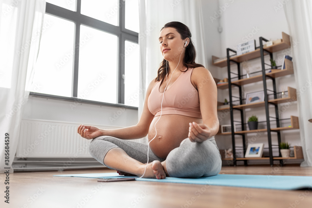 sport, yoga and people concept - happy pregnant woman with earphones and smartphone listening to music and meditating at home
