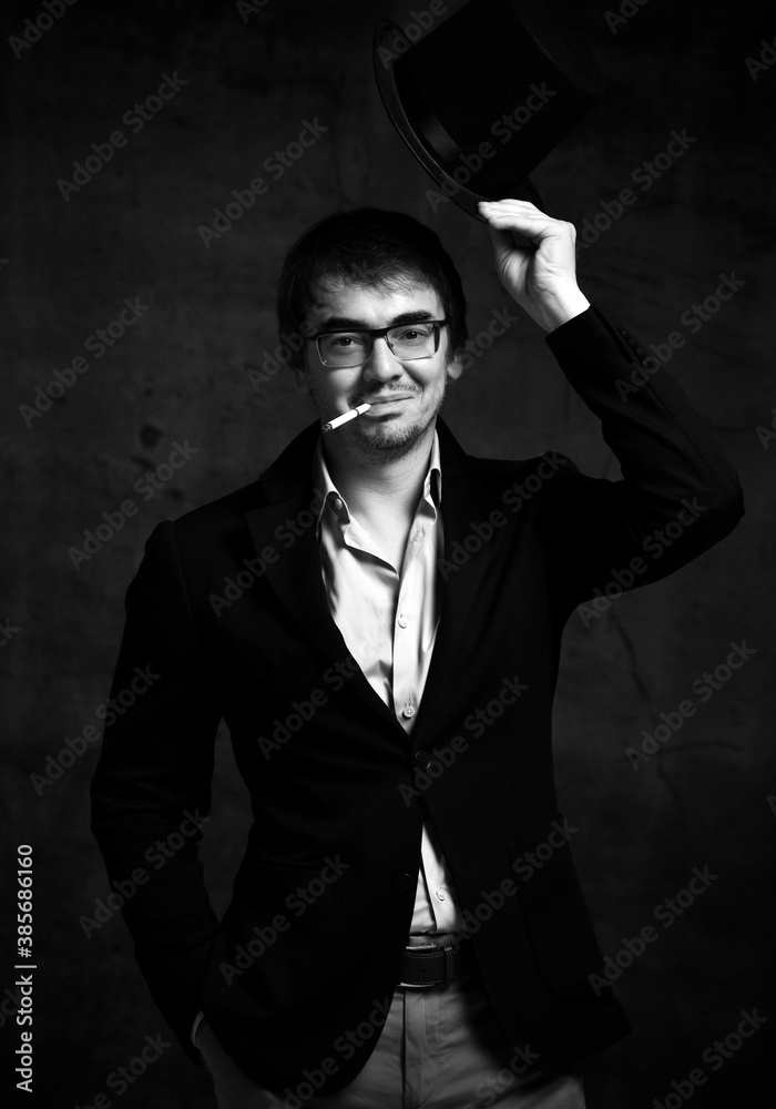 Smoking cigarette unshaved friendly man in black costume and glasses holds hat in raised hand greeting Smoking cigarette unshaved friendly man in black costume and glasses holds hat in raised hand greeting