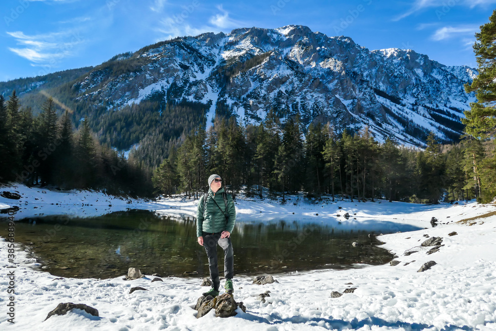 A man standing at the stone next to the shore of Green Lake in Austrian Alps. Mountains and ground are covered with snow. Winter hiking. The man is enjoying a frosty winter day. Winter wonderland A man standing at the stone next to the shore of Green Lake in Austrian Alps. Mountains and ground are covered with snow. Winter hiking. The man is enjoying a frosty winter day. Winter wonderland