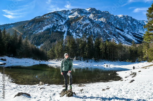 A man standing at the stone next to the shore of Green Lake in Austrian Alps. Mountains and ground are covered with snow. Winter hiking. The man is enjoying a frosty winter day. Winter wonderland