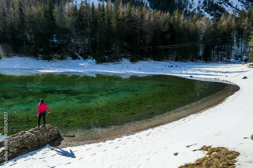 A woman standing at the stone next to the shore of Green Lake in Austrian Alps. Mountains and ground are covered with snow. Winter hiking. She is enjoying a frosty winter day. Winter wonderland