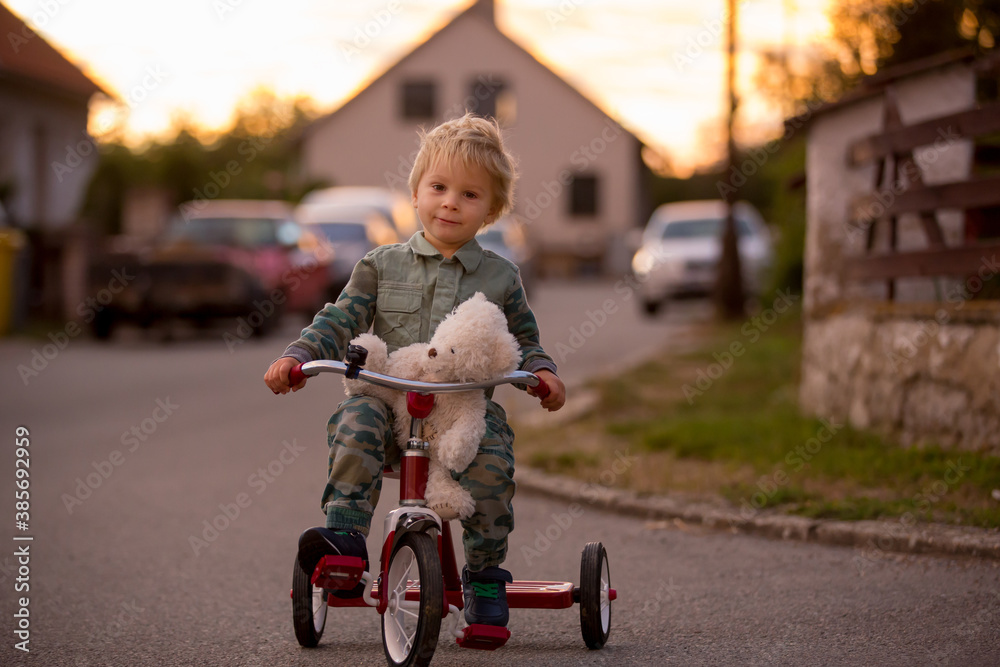 Toddler child, blond boy, riding tricycle in a village small road on sunset Toddler child, blond boy, riding tricycle in a village small road on sunset