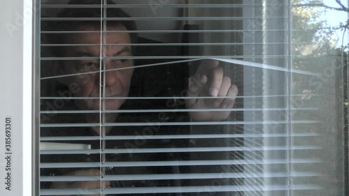 A man is observing with binoculars through the blinds in the window.