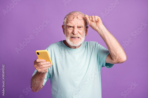 Photo portrait of cranky old man lifting up glasses holding phone in one hand isolated on vivid violet colored background